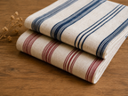 Two folded striped fabric napkins or kitchen towels stacked on a wooden surface. The top towel is cream with dark blue stripes, and the bottom towel is cream with red stripes.