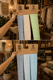 A person in a rustic boutique holds up a cardboard hanger displaying three fabric swatches: light blue, cream, and sage green. The bottom half shows the same fabrics folded and arranged on a table in the shop.